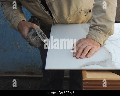 a craftsman with a grinder sharpens the edge of a large ceramic tile in an outdoor workshop, processing facing tiles with a round sharp disc Stock Photo