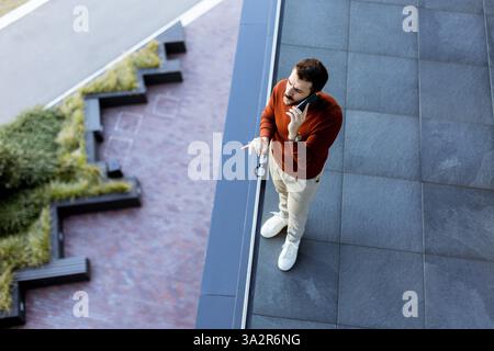 A young man stands on a sleek balcony while engaged in an animated phone conversation. Below him, a blend of contemporary design and lush greenery enh Stock Photo