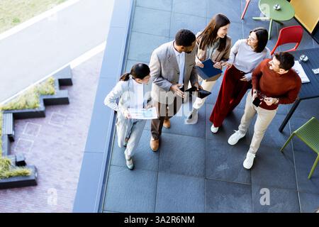 A group of five professionals gathers on an outdoor terrace, sharing ideas and plans. They hold various devices and papers, showcasing a collaborative Stock Photo