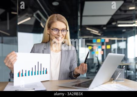Professional woman presenting a bar chart via video conference from her office, using a laptop. She is wearing a grey blazer and glasses. Stock Photo