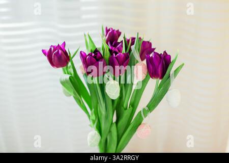 Easter home decor. Eggs hung on purple tulips in vase. Bouquet of spring flowers blooming in living room Stock Photo