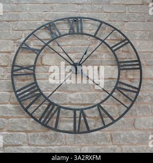 A large metal wire frame clock with roman numerals hangs on a grey stone wall. Stock Photo