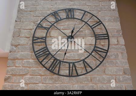 A large metal wire frame clock with roman numerals hangs on a grey stone wall. Stock Photo