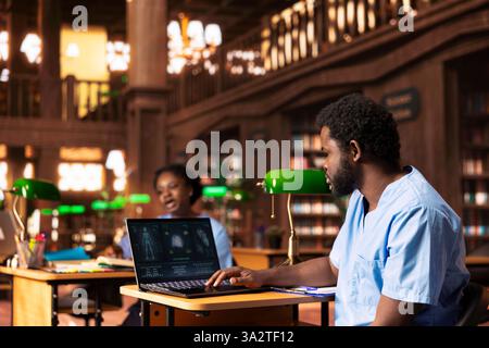 African american med school pupil studies pharmacology notes in a library, preparing for exams and advancing their healthcare career. Man in scrubs works hard on medical school success. Stock Photo