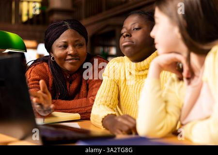 Preteen scholars collaborating with their tutor in a learning environment, working on school projects and developing critical classroom skills. Woman mentoring her students in the library. Stock Photo