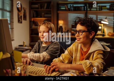 Two boys smiling while using computer in warmly lit room filled with books and personal items around desk, creating cozy atmosphere Stock Photo