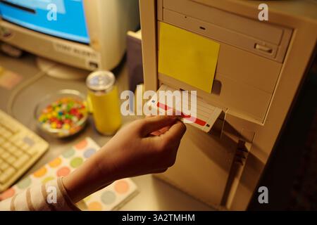 Hand inserting floppy disk into old desktop computer tower with yellow note and candies nearby in soft light. Retro setup featuring vintage technology and personal touch Stock Photo