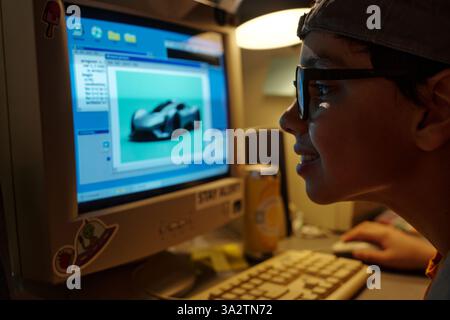 Boy wearing glasses and cap smiling while working on computer. Screen displaying automotive design software creating innovative project featuring futuristic car model Stock Photo