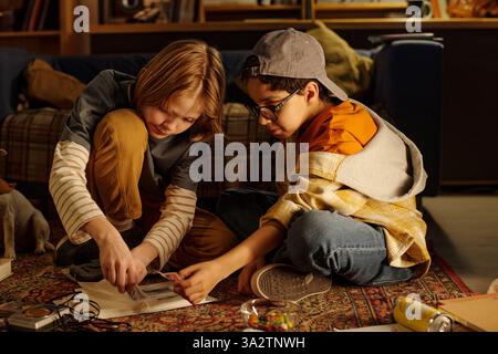 Children sitting on floor engaged in hands-on creative activity, focused on working together. Multiracial children enjoying project on patterned rug in cozy home setting Stock Photo