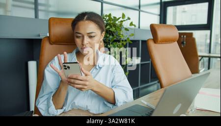 A young woman sitting in a modern office checking her smartphone, surrounded by plants, leather chairs, and a laptop on a clean and stylish desk Stock Photo