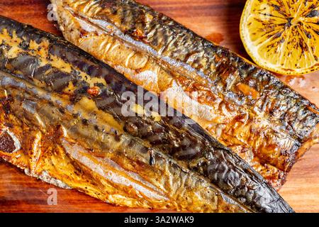 Two grilled mackerel fillets are placed on a wooden board. Accompanying the fish is a half lemon, lightly charred, adding a hint of citrus. The dish l Stock Photo
