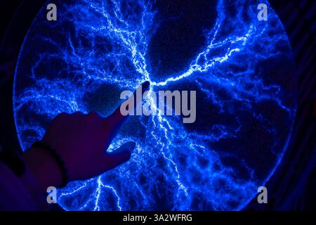 Hand touching a plasma display, creating mesmerizing blue electrical discharges in a dark environment, showcasing the fascinating interaction between Stock Photo