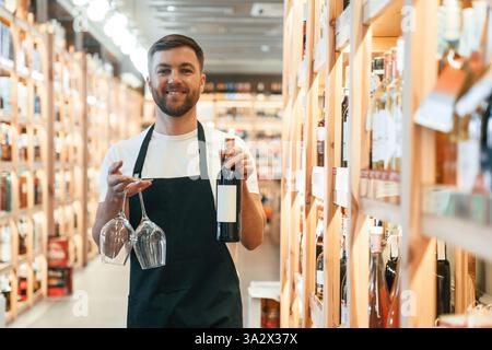 Two glasses and bottle in hands. Wine shop owner in white shirt and black apron. Stock Photo