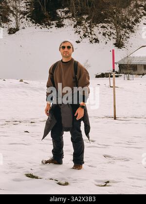 A man wearing a brown sweater and black pants stands in the snow. He is wearing a backpack and a watch Stock Photo