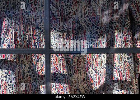 Viewed through a window, two intersecting metal bars form a tilted cross against a flowing dress with red and blue abstract patterns on a white canvas Stock Photo