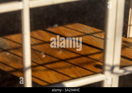 Through a window with square wooden grates, shadows align perfectly on the worn wooden floor, creating a rhythmic pattern that echoes the window Stock Photo
