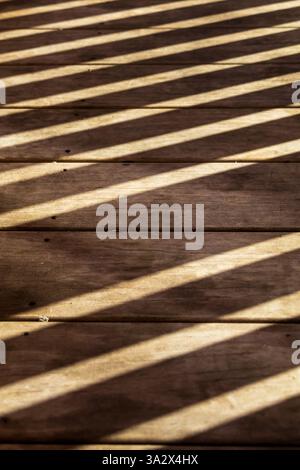 Diagonal, parallel shadows from a railing fall across an outdoor wooden floor, where smooth planks run horizontally in perfect alignment with the shot Stock Photo