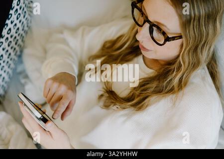 Mother in her 40s relaxing in bed, scrolling on phone in a light room Stock Photo