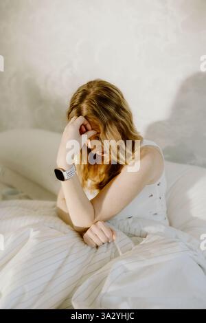 A tired woman sits in bed, holding her head in a moment of stress Stock Photo