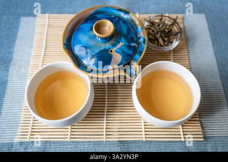 Two white cup of tea, nice blue ceramic kettle, dried loose tea leaves in small glass bowl. All on gentle bamboo pad on blue background. Stock Photo