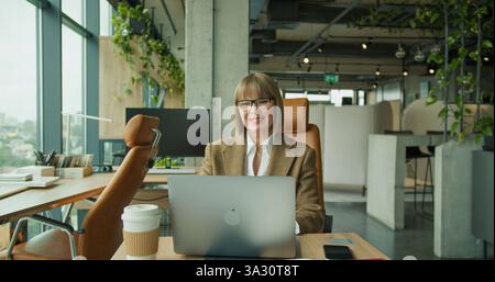 A senior professional woman working on a laptop in a modern office setting with large windows and lush greenery, symbolizing efficiency and Stock Photo