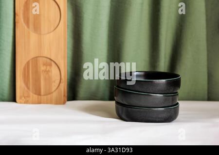 Minimalist composition with stacked black bowls, a wooden tray, and a green fabric backdrop Stock Photo