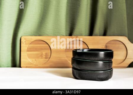 Minimalist composition with stacked black bowls, a wooden tray, and a green fabric backdrop Stock Photo