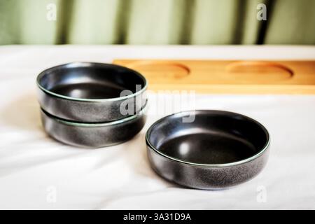 Black ceramic bowls arranged on a white tablecloth with a wooden tray and green fabric in the background Stock Photo