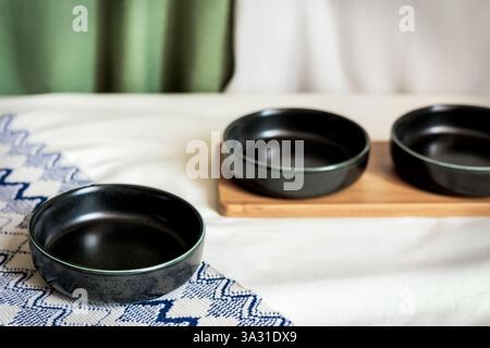 Black ceramic bowls on a white tablecloth with a blue embroidered napkin and wooden tray, creating a cozy and elegant dining setup Stock Photo