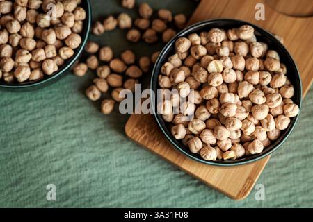 A ceramic bowl filled with dried chickpeas placed on a wooden tray, surrounded by scattered legumes on a green textured background, showcasing a rusti Stock Photo