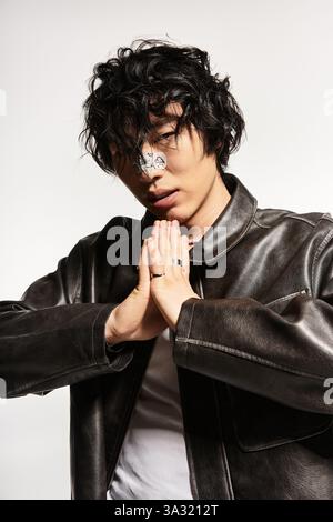 Stylish Asian man with plaster on nose posing in leather jacket with clasped hands in studio. Stock Photo
