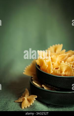 Close-up of uncooked farfalle pasta in stacked black ceramic bowls on a green fabric background. Minimalist and rustic composition, ideal for culinary Stock Photo