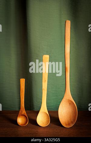Three wooden spoons of different sizes standing upright on a wooden surface against a green fabric background. Minimalist and rustic kitchen compositi Stock Photo