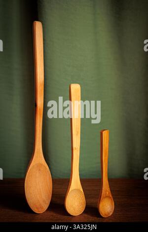 Vertical arrangement of three wooden spoons of varying sizes placed on a wooden surface with a green fabric backdrop. Simple and rustic setup, perfect Stock Photo