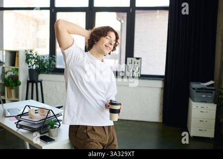Handsome young man smiles and stretches at his desk during a refreshing coffee break in the office. Stock Photo