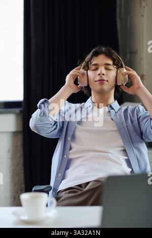 Man with curly hair wearing headphones relaxes at his desk, savoring a moment of calm and focus. Stock Photo