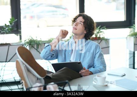 A young handsome man relaxes in an office, gazing thoughtfully while holding a tablet. Stock Photo