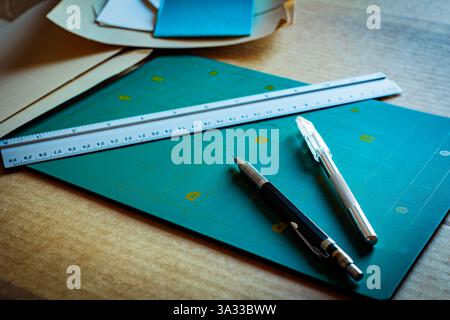 Workspace with crafting tools, including a cutting mat, precision knives, and a metal ruler, arranged with paper sheets on a wooden surface. Perfect f Stock Photo