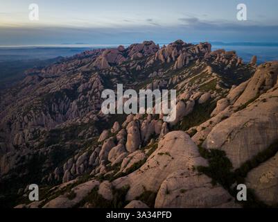 Aerial view of dramatic rock formations in Montserrat, Catalonia, at sunset. The rugged landscape reveals reddish stone peaks contrasted against a ser Stock Photo