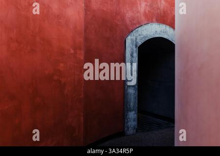 This image features a minimalist architectural design with a striking contrast between vibrant red walls and a weathered gray arched doorway. The simp Stock Photo