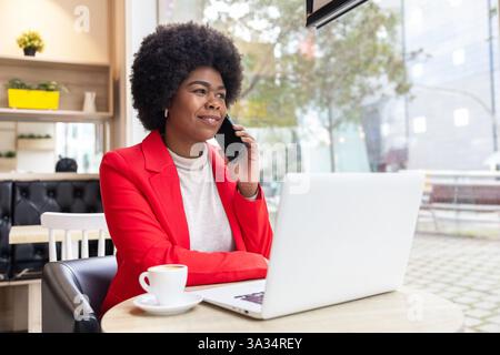 Confident woman in a vibrant red blazer talks on her phone while working on a laptop in a bright, cozy cafe. Stock Photo