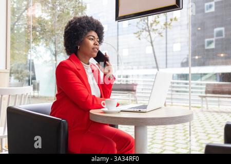 A professional woman in a red suit sits in a modern cafe, talking on her phone while working on a laptop. This image captures a dynamic urban lifestyl Stock Photo