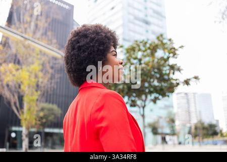 A confident woman wearing a red blazer stands in an urban setting with skyscrapers in the background. Her curly hair and calm expression highlight her Stock Photo