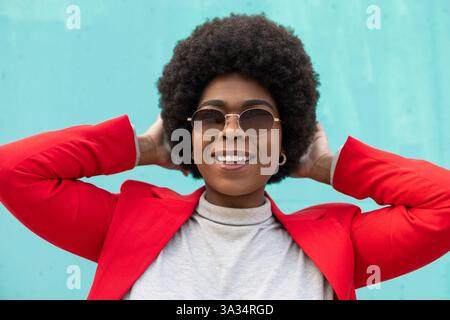 A smiling woman in stylish sunglasses and a vibrant red jacket confidently poses with her hands on her head against a turquoise backdrop, exuding a ch Stock Photo