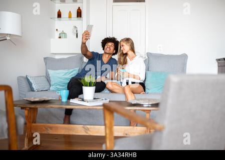 A young couple spends a relaxing day in a bright living room, sitting on a modern sofa. They enjoy taking a selfie together, capturing moments on a su Stock Photo