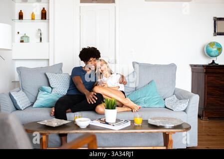 A happy couple sits on a cozy couch, looking at a phone. The bright living room has modern decor, with a globe on a nearby table, creating a relaxed a Stock Photo