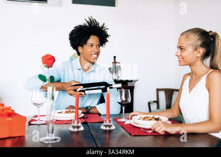 A man and woman share a romantic dinner at home, featuring wine, red candles, and a rose. The intimate setting highlights their connection and joyful Stock Photo