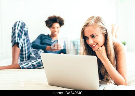 A young woman smiles while using a laptop on her bed, with a man in the background holding a phone, enjoying a relaxed day indoors with technology and Stock Photo