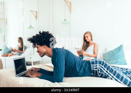 A young man using a laptop and a woman browsing a tablet in a cozy bedroom. They are casually dressed in leisurewear, enjoying a relaxed morning at ho Stock Photo
