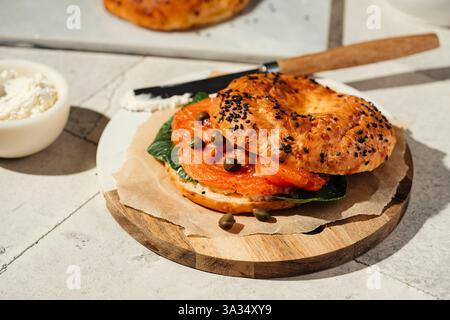 A freshly baked sesame bagel filled with smoked salmon, capers, and greens on a wooden plate. Light cream cheese is spread with a knife, adding a crea Stock Photo